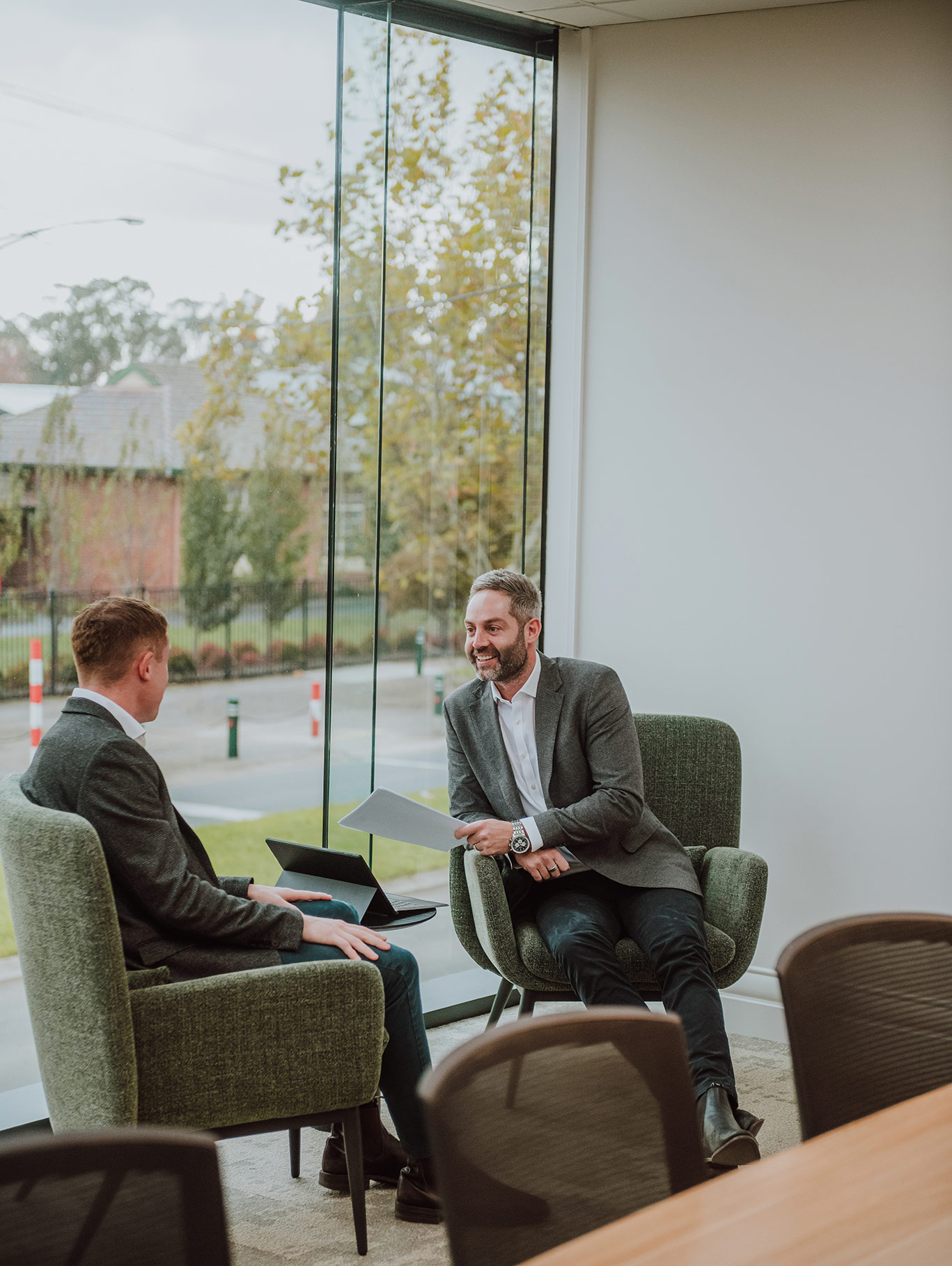 Photo of two business men in shirts, jacket and jeans, sitting in green armchairs in a modern office meeting space with large windows behind them.