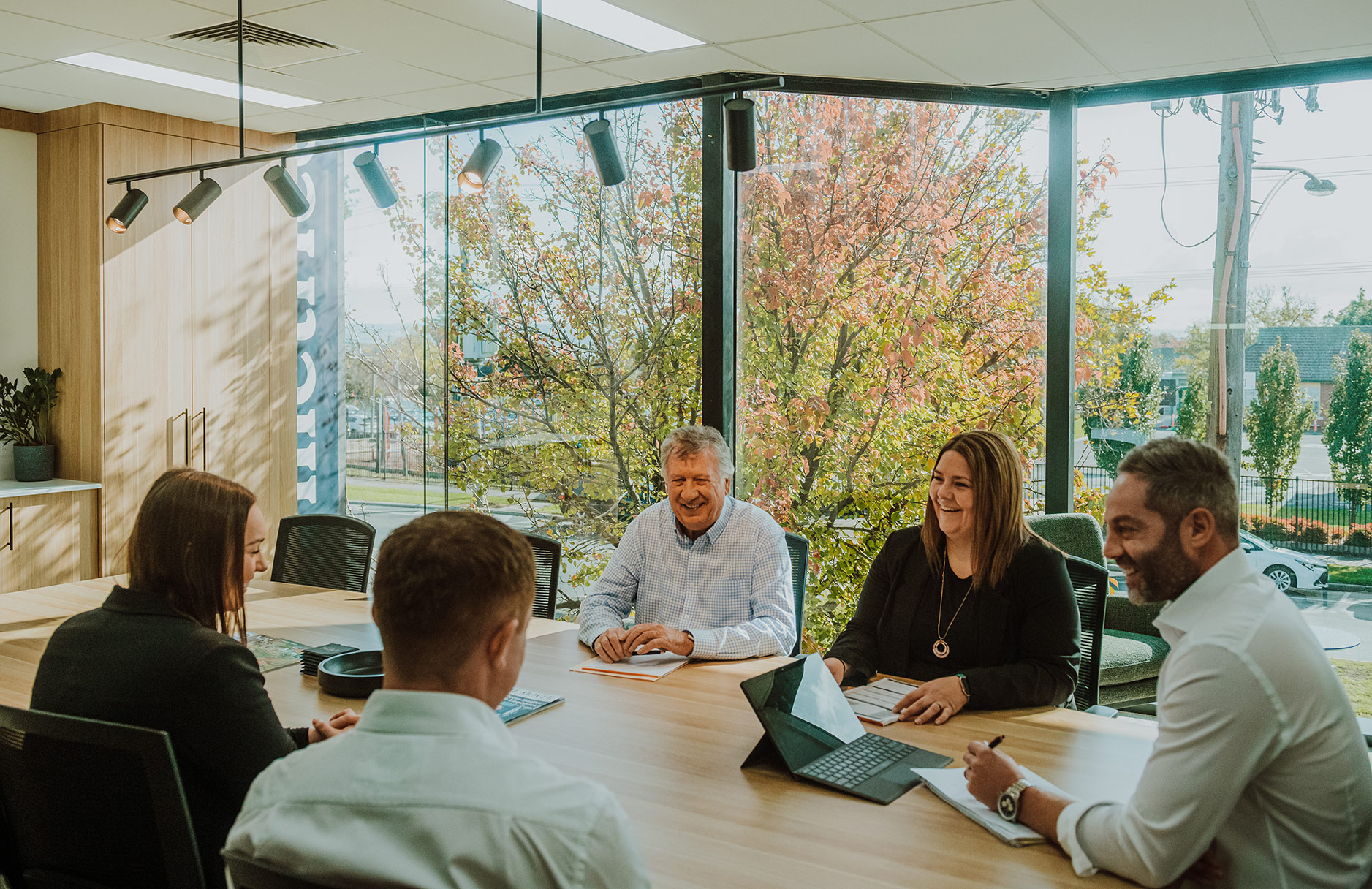 Photo of a business team of three men and two women sitting at a table in a contemporary meeting room with large windows behind them.