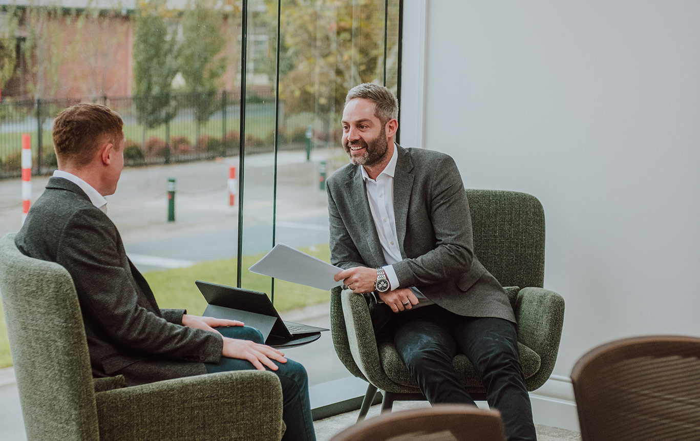 Photo of two business men in shirts, jacket and jeans, sitting in green armchairs in a modern office meeting space with large windows behind them.