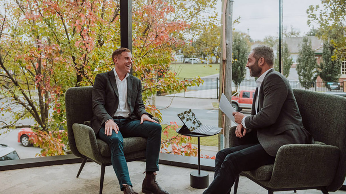 Photo of two business men in shirts, jacket and jeans, sitting in green armchairs in a modern office meeting space with large windows behind them.
